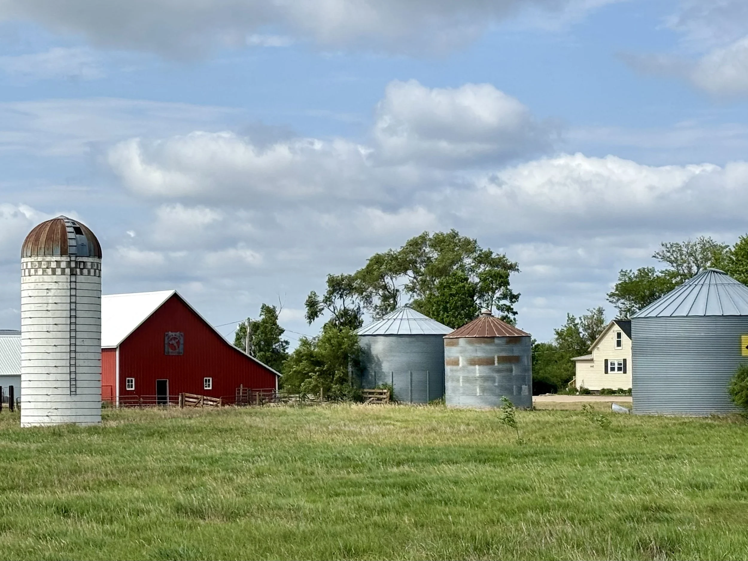 A farm scene with a red barn, several metal silos, and a white house, surrounded by green grass and trees under a partly cloudy sky.