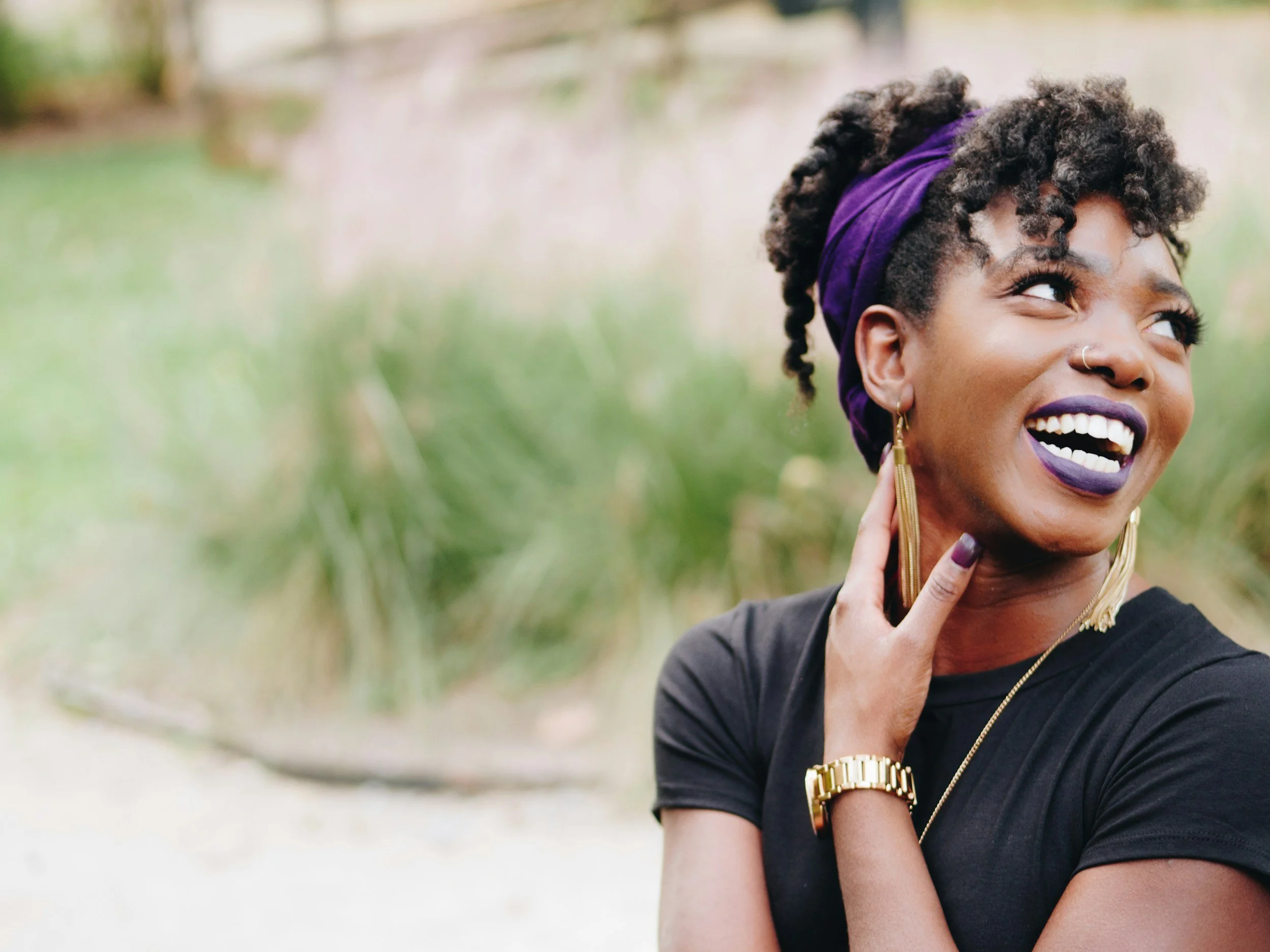 A smiling woman with dark curly hair wearing a purple headband, gold jewelry, and a black top, outdoors in a natural setting.