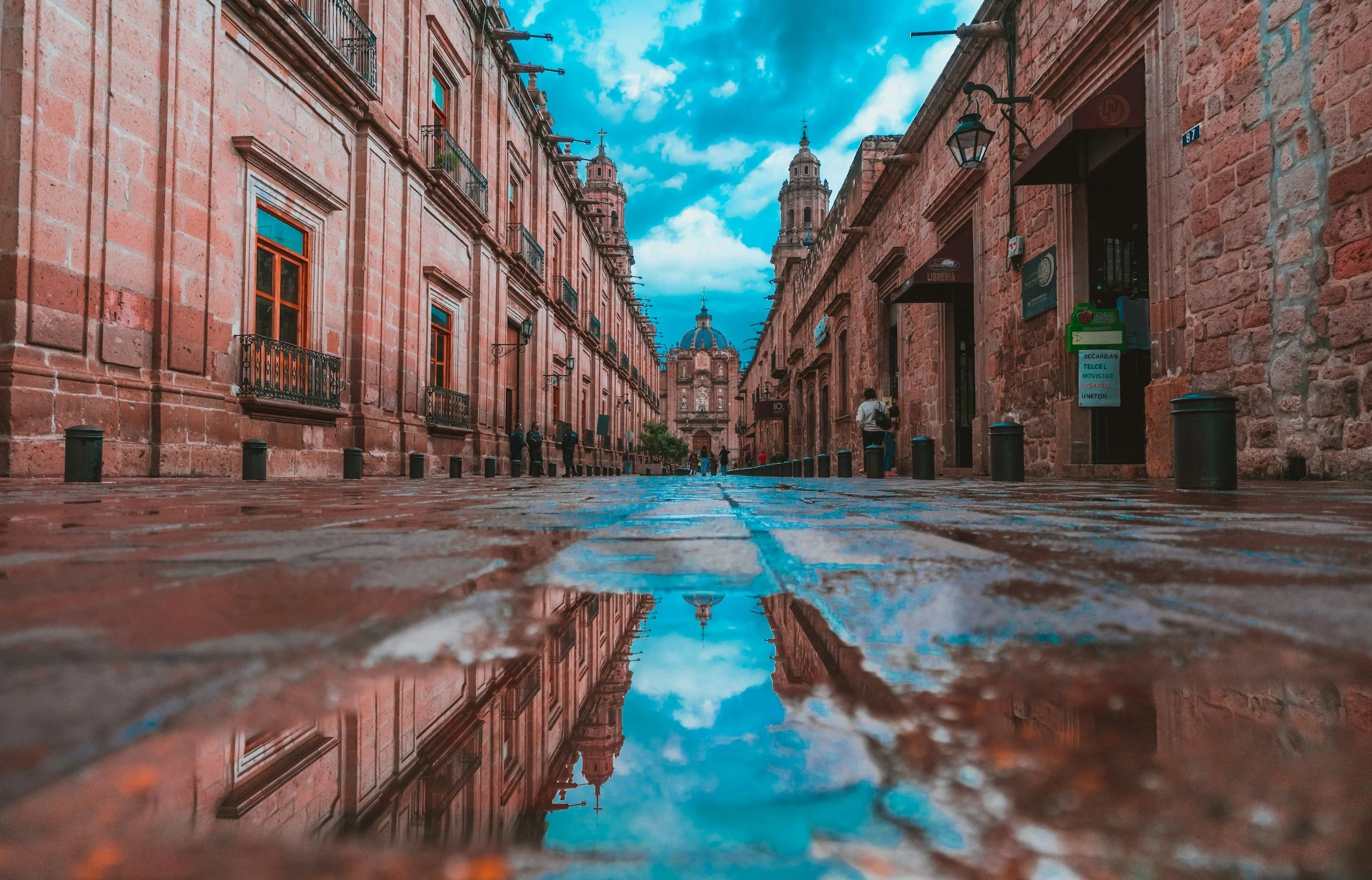 A cobblestone street lined with historic architecture and domed cathedral in the distance, reflected in a puddle.
