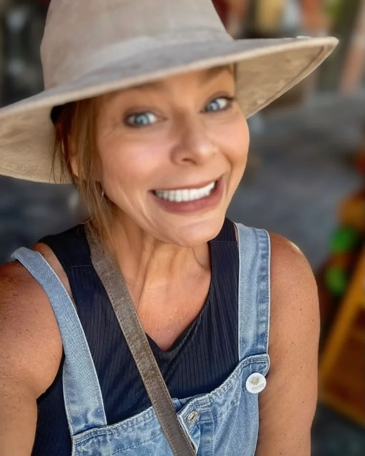 A woman with short reddish hair wearing a wide-brimmed hat, a black tank top, and denim overalls, smiling at the camera.