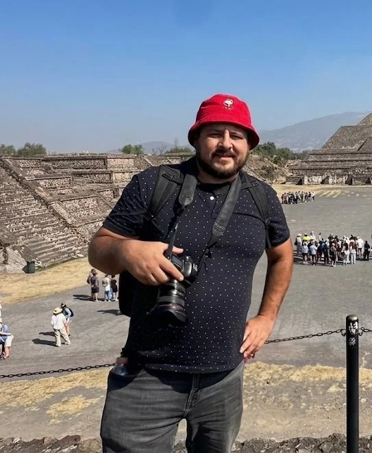 A man wearing a red bucket hat, black dotted shirt, and carrying a camera and backpack standing at an ancient archaeological site with stepped stone structures and a group of tourists in the background.
