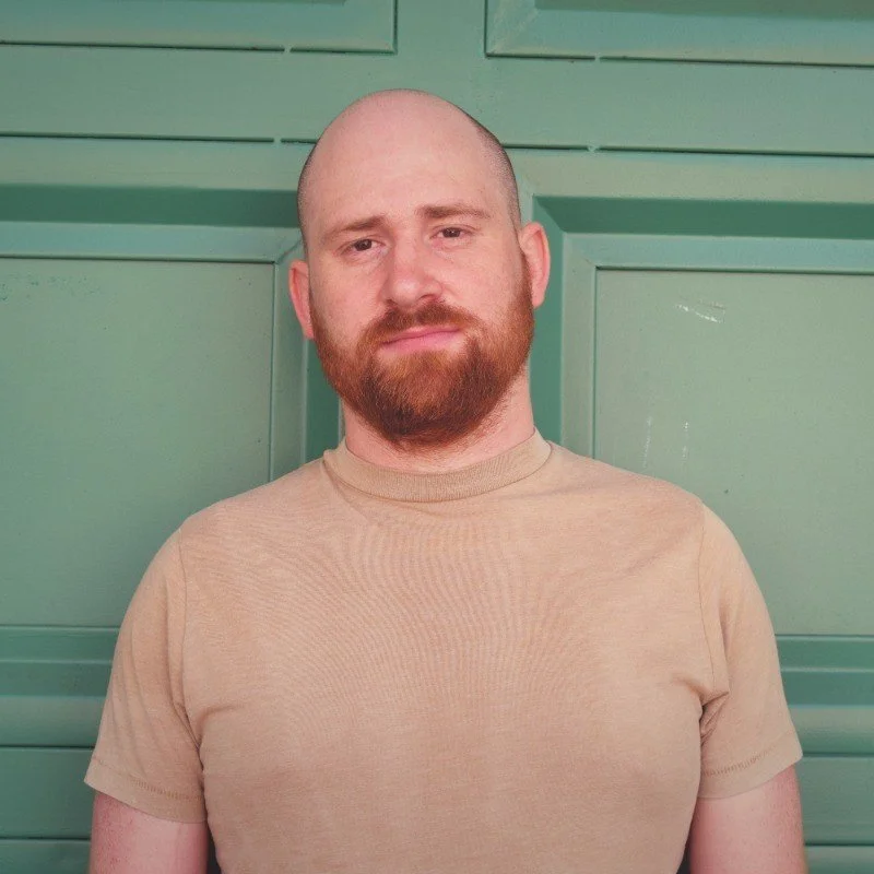 A man with a shaved head and a beard, wearing a beige T-shirt, standing in front of a green door.