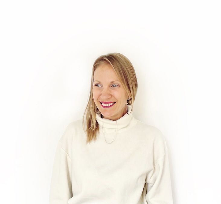 A young woman with blonde hair smiling and looking to the side, wearing a white turtleneck sweater and earrings, against a plain white background.