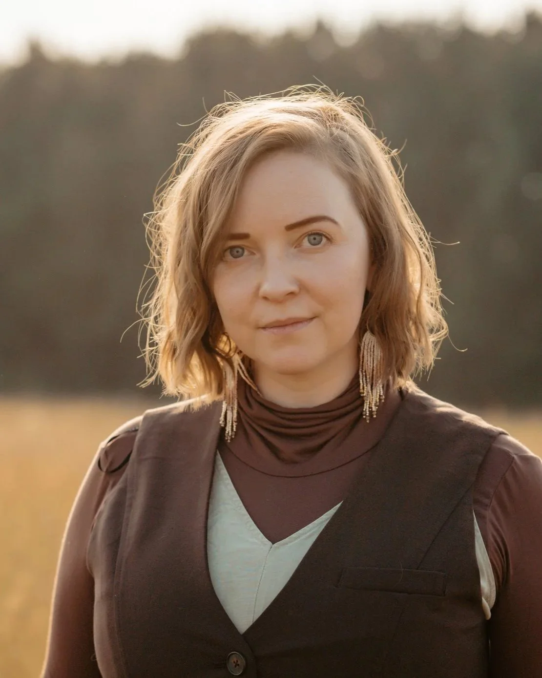 A woman with shoulder-length wavy blonde hair and blue eyes standing outdoors in a field during sunset, wearing a brown vest over a cream top with long earrings.