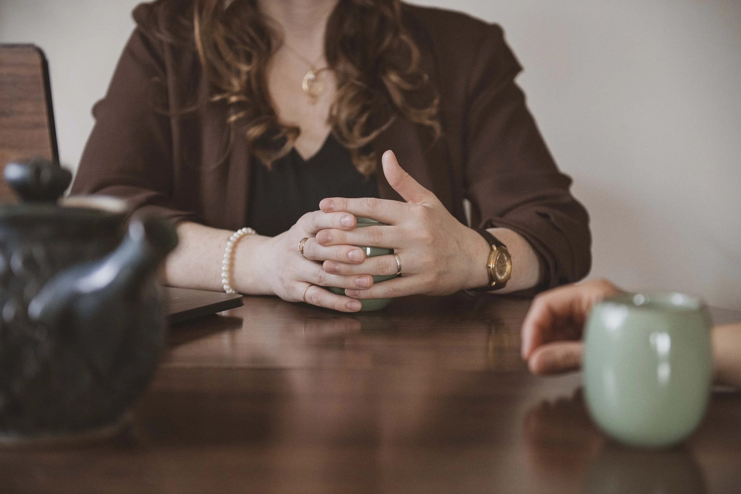 A woman with wavy, reddish-brown hair sitting at a wooden table with her hands clasped on a green mug. She is wearing a dark top, a brown blazer, and accessories including a watch, rings, and a pearl bracelet. Part of a camera and another green mug are also visible on the table.