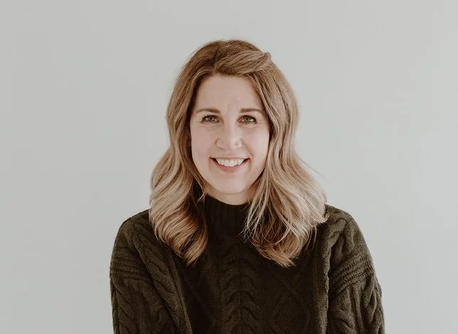 A woman with wavy, shoulder-length blonde hair, wearing a black sweater, smiling at the camera against a plain light background.