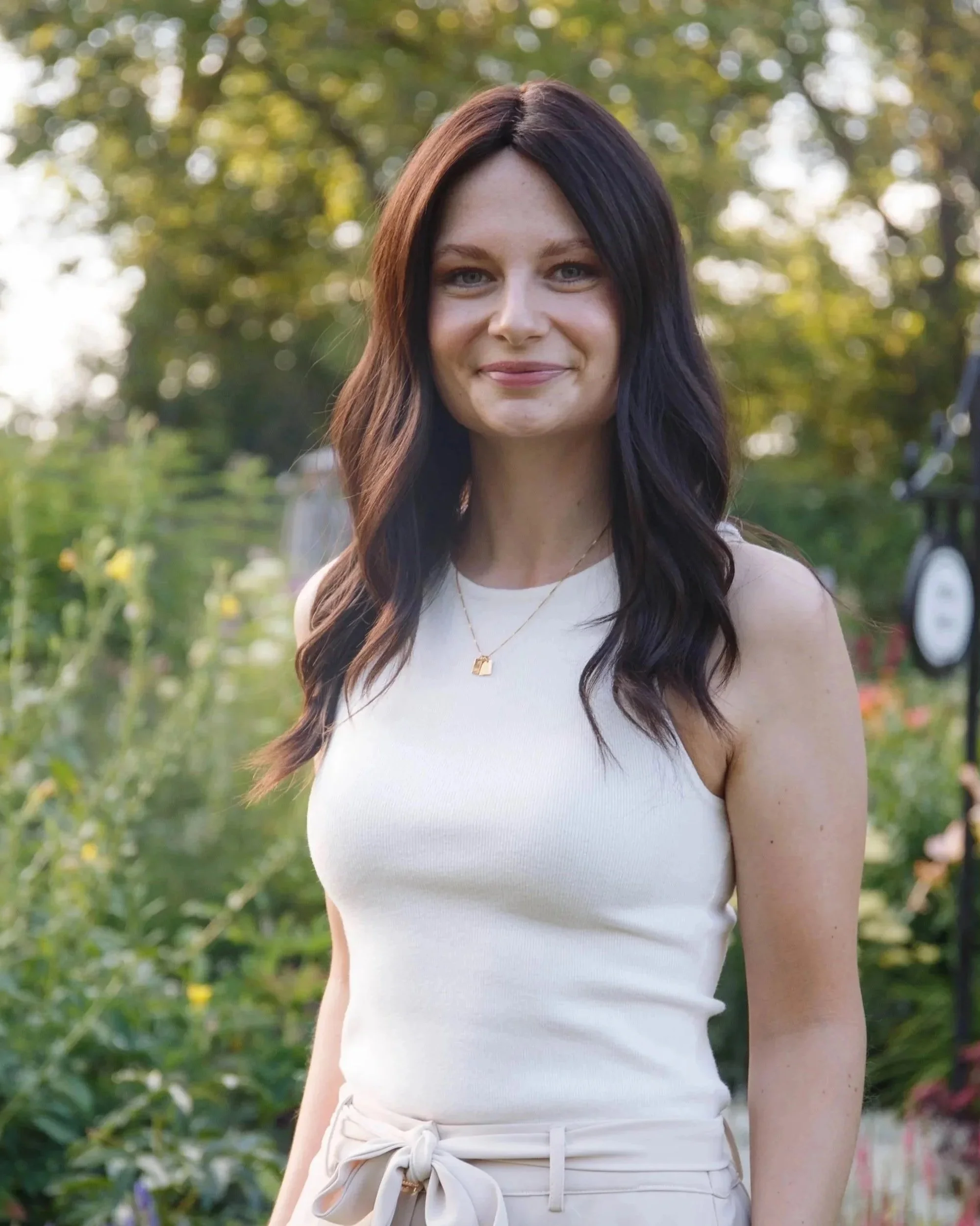 A woman with long dark hair smiling outdoors, wearing a white sleeveless top and a gold necklace, with trees and greenery in the background.