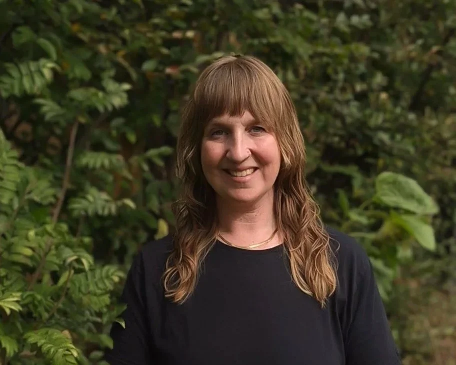 A woman with shoulder-length wavy brown hair, smiling, wearing a black top, standing outdoors in front of green foliage.