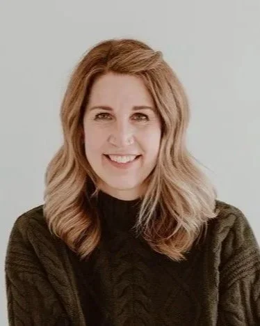 A woman with wavy, shoulder-length light brown hair, wearing a dark sweater, smiling against a light gray background.