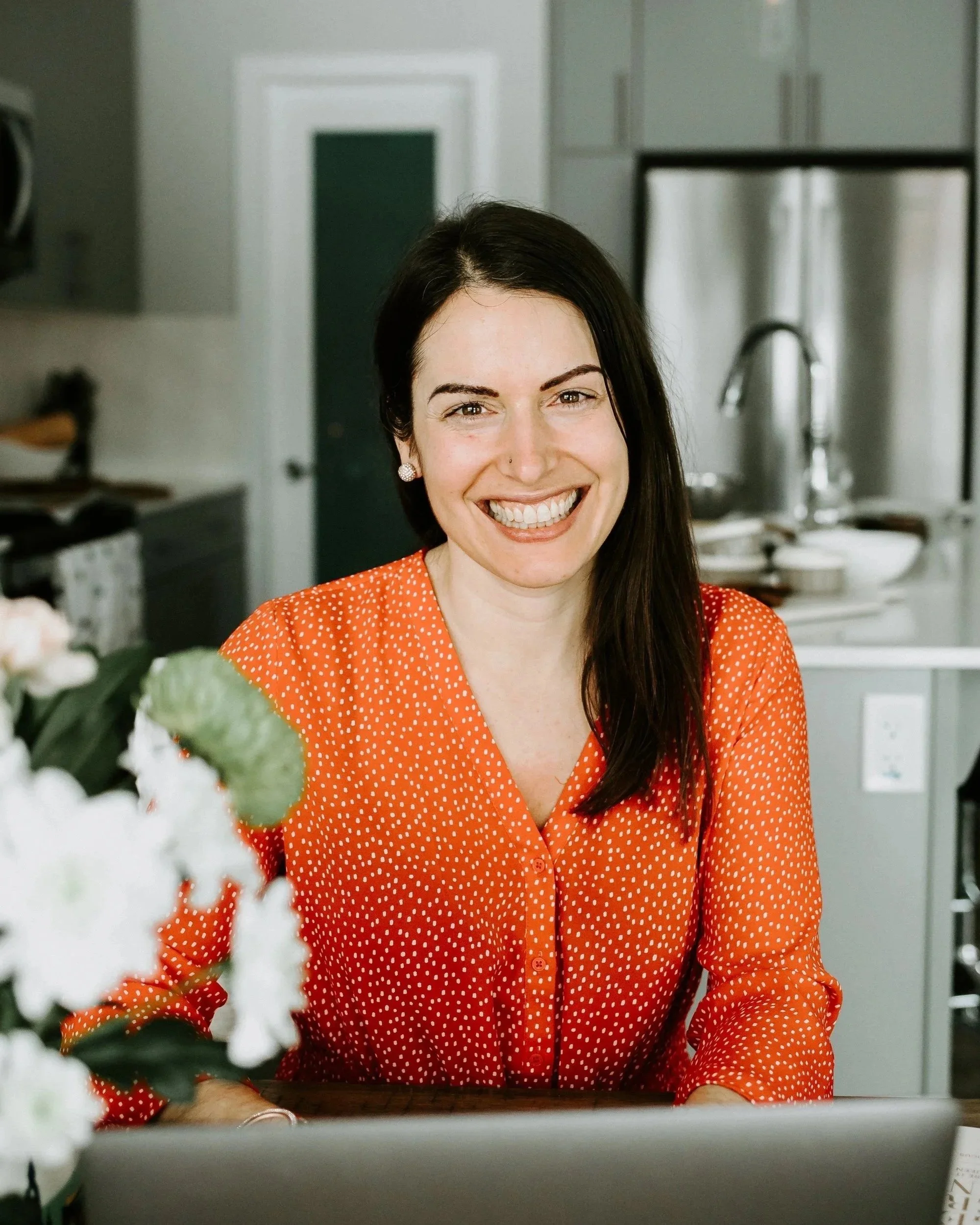 Woman with dark hair and a wide smile wearing a red dress with white polka dots, sitting at a table in a kitchen.