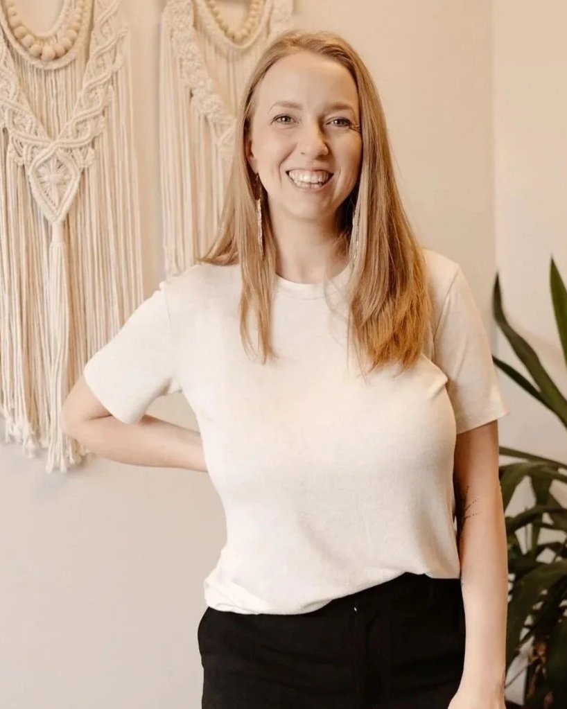 A woman with long red hair wearing a white t-shirt and black pants, standing indoors in front of a wall hanging and a plant, smiling at the camera.
