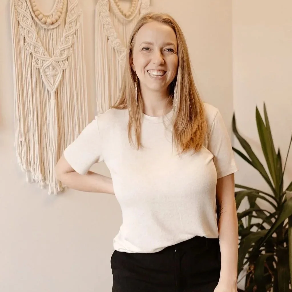 A woman with long red hair smiling and winking, wearing a white t-shirt and black pants, standing indoors against a wall with macramé wall hanging and next to a large leafy plant.