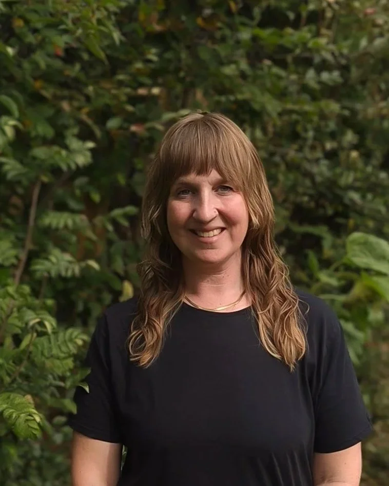 Smiling woman with wavy light brown hair wearing a black t-shirt, standing outdoors in front of lush green foliage.