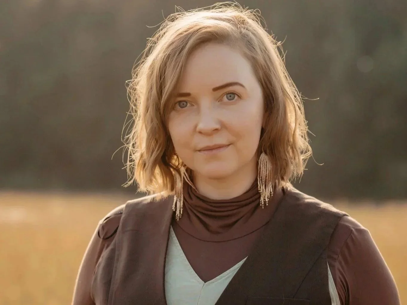 A woman with short, wavy, light brown hair and blue eyes stands outdoors in a field during sunset, wearing earrings and a brown vest over a white top.