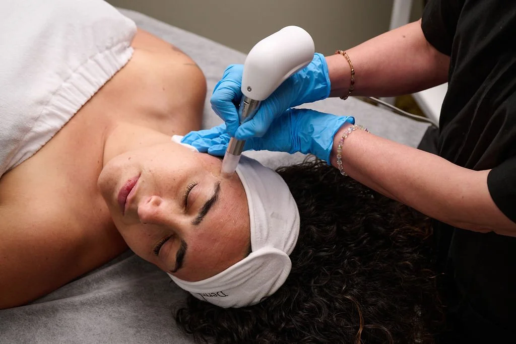 A woman lying face up with a cloth headband, receiving a facial treatment using a handheld device by an esthetician wearing blue gloves.