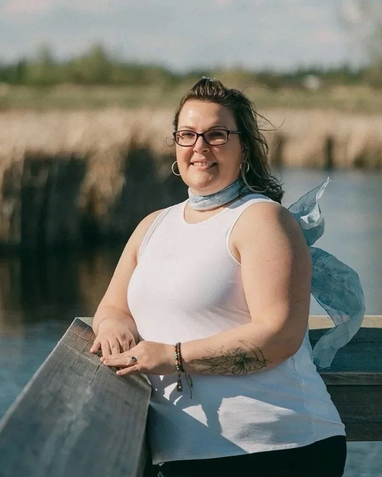 A woman smiling outdoors near water, wearing glasses, a white sleeveless top, and a blue scarf around her neck, with her arm resting on a wooden railing.
