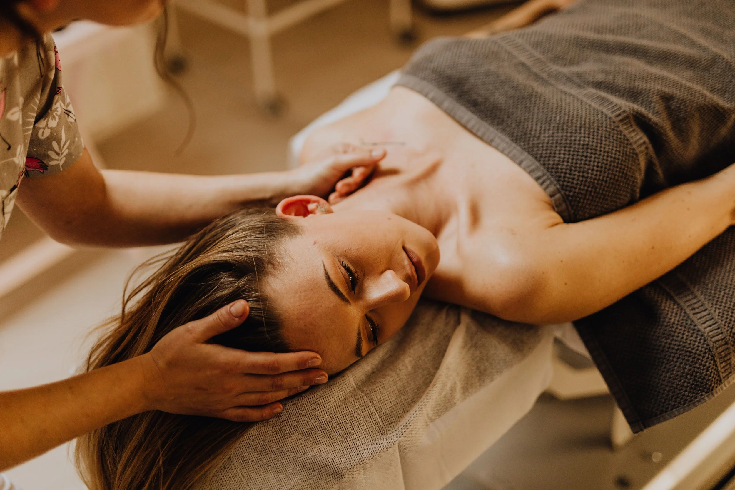 A woman lies face down on a massage table with her eyes closed, receiving a head massage. A massage therapist's hands gently support her head and neck. The woman appears relaxed, with long hair spread out on the table.