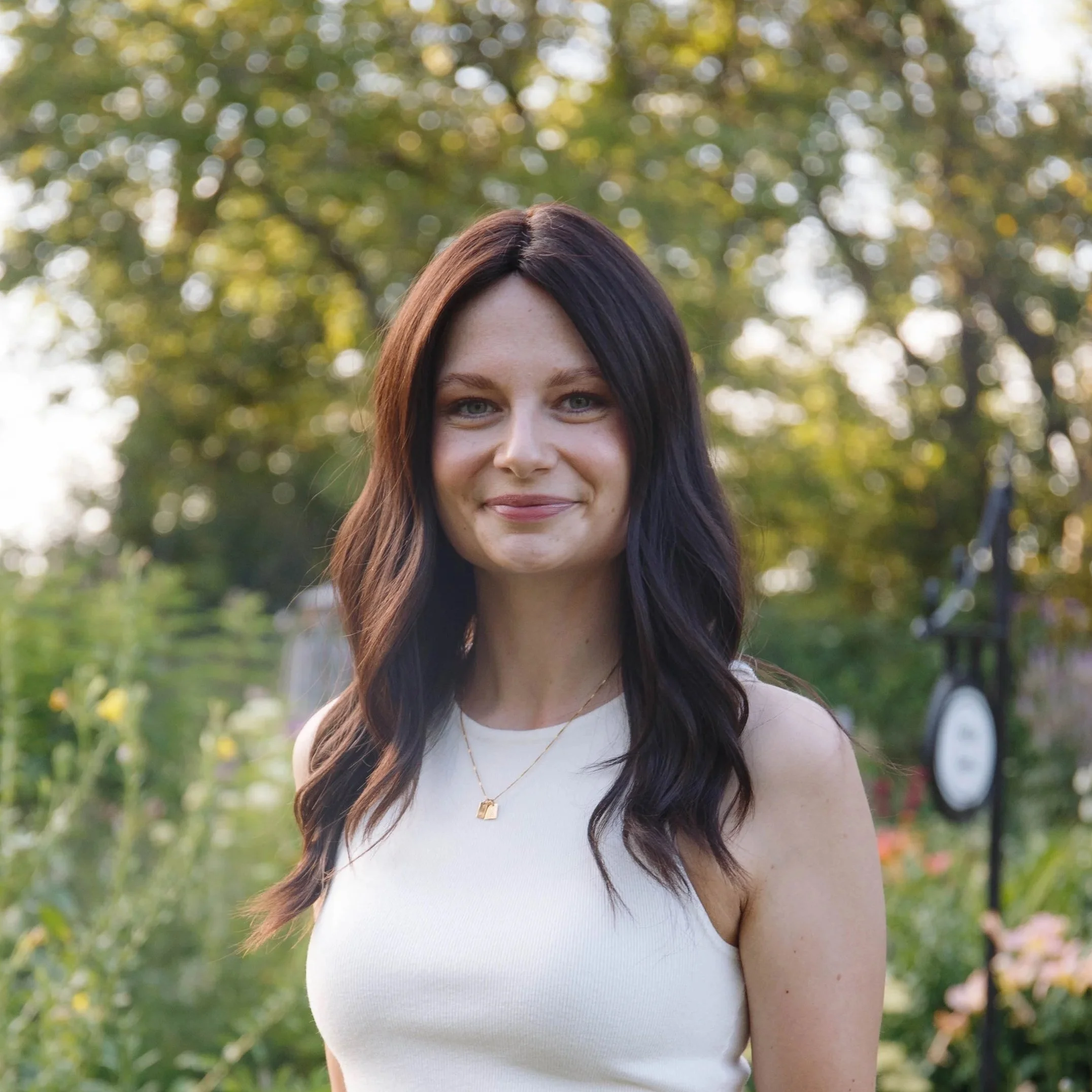 A woman with long dark brown hair and blue eyes smiling, wearing a white sleeveless top and a gold necklace, standing outdoors with a blurred background of green trees and sunlight.