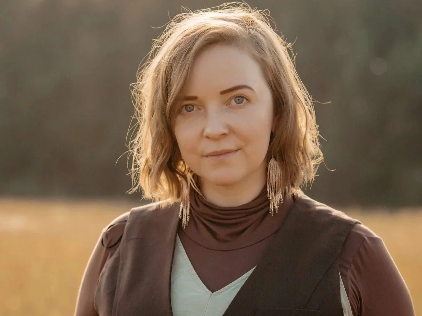 A woman with shoulder-length wavy blonde hair, wearing a brown sleeveless vest over a white shirt, and long earrings, standing outdoors in a sunny setting with blurred background.
