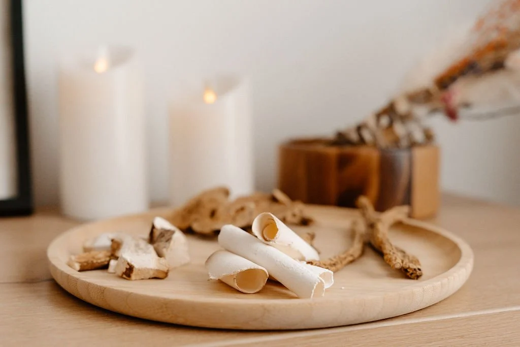 Wooden tray with rolled white cookies and broken pieces, on a light wood surface, with white candles, a wooden container, and decorative branches in the background.