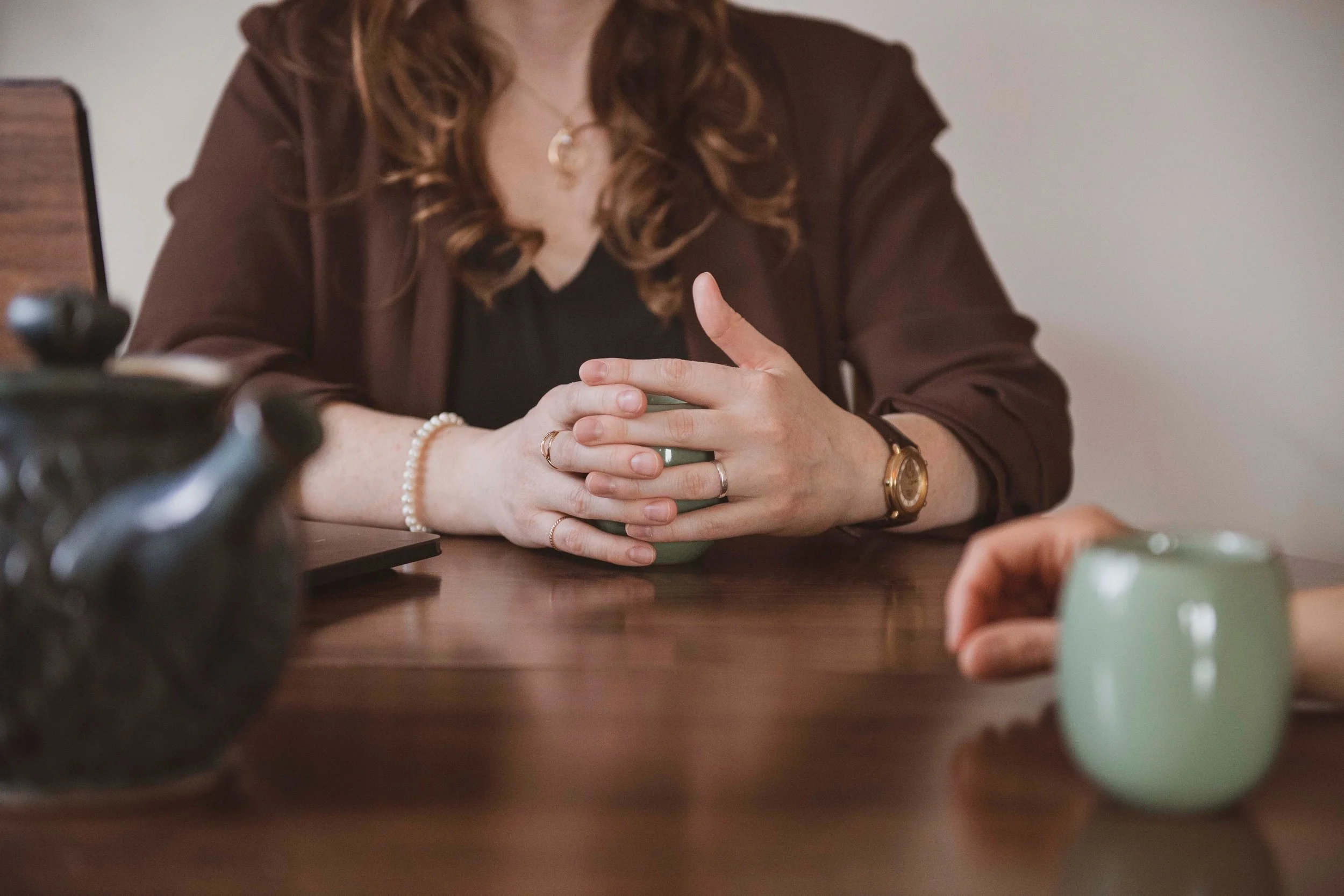 A woman with curl hair, wearing a dark jacket and jewelry, sitting at a table with her hands clasped around a mug. Part of her face is not visible in the frame.