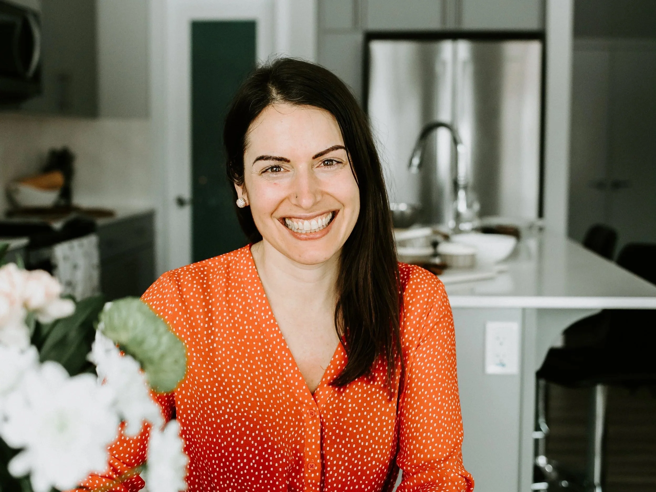 Close-up of a woman with long dark hair, wearing an orange dress with white polka dots, smiling in a kitchen.