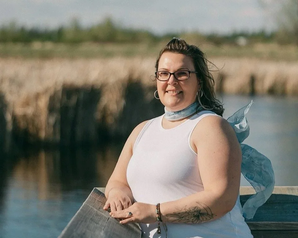 A woman with glasses, earrings, and tattoos is smiling while standing on a boat near a body of water, with fields in the background.