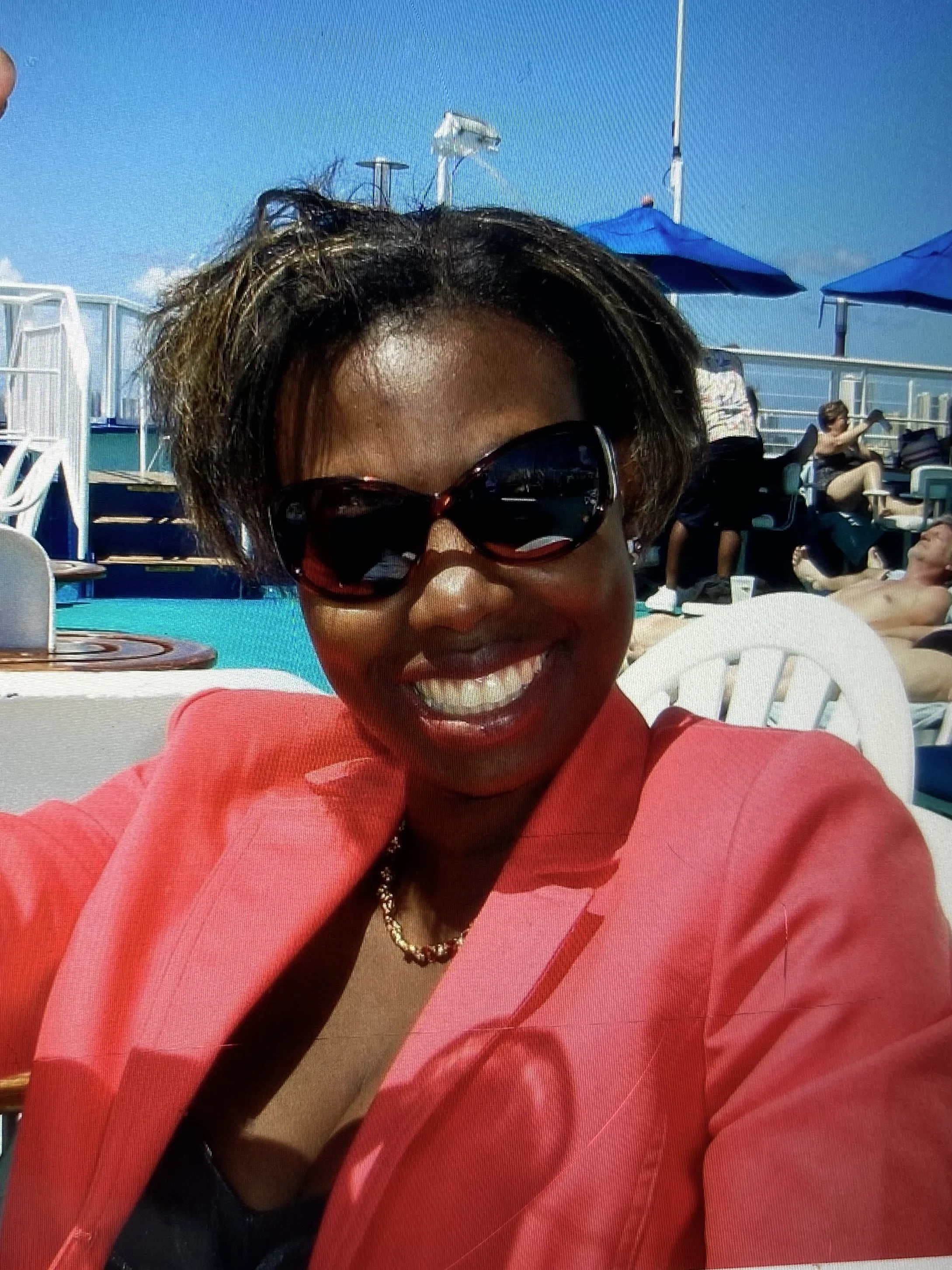 A woman smiling, wearing large sunglasses and a pink jacket, sitting outdoors with a pool and blue umbrellas in the background.