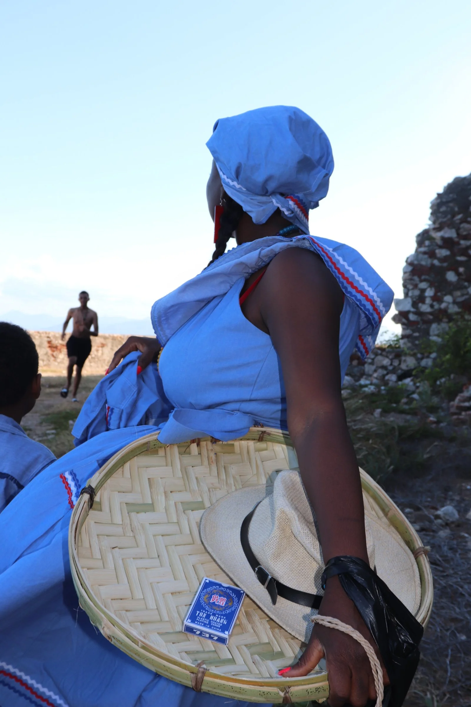 A woman wearing a blue headscarf and matching dress holding a woven basket with a hat and a pack of Marlboro cigarettes inside. She is outdoors with a man running in the background and rocks on the side.