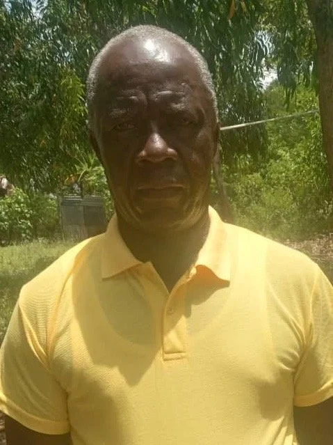 A middle-aged man with dark skin and short hair, wearing a yellow polo shirt, standing outdoors with green trees and sunlight in the background.