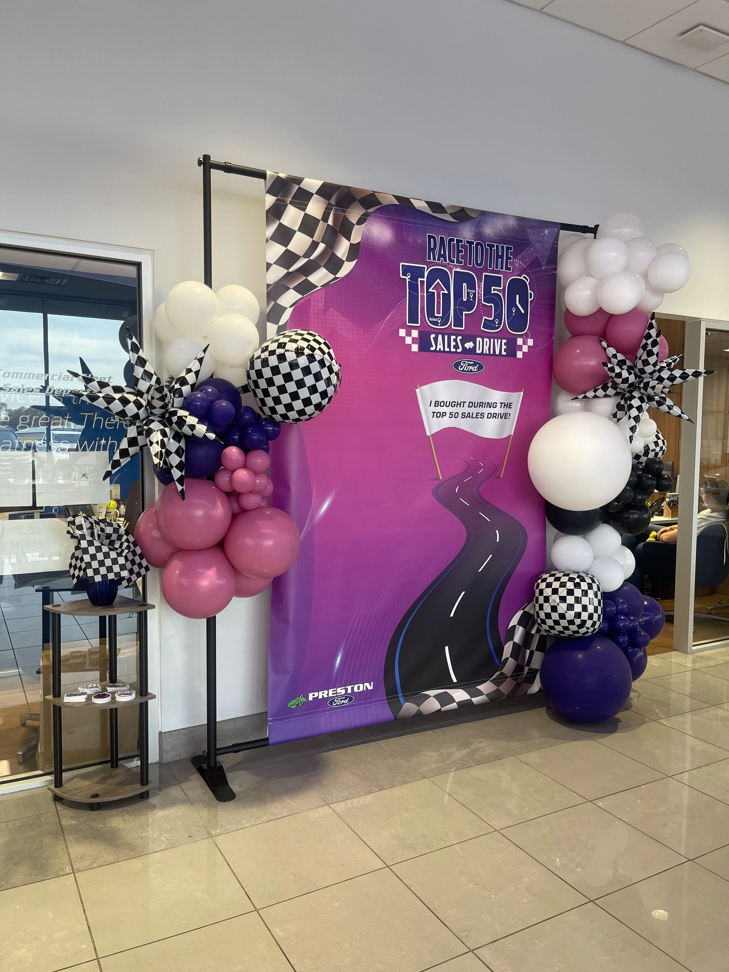 Decorative display for a sales event with balloons in purple, black, white, and pink, a banner with checkered flags and a racing theme, and a small table with accessories.