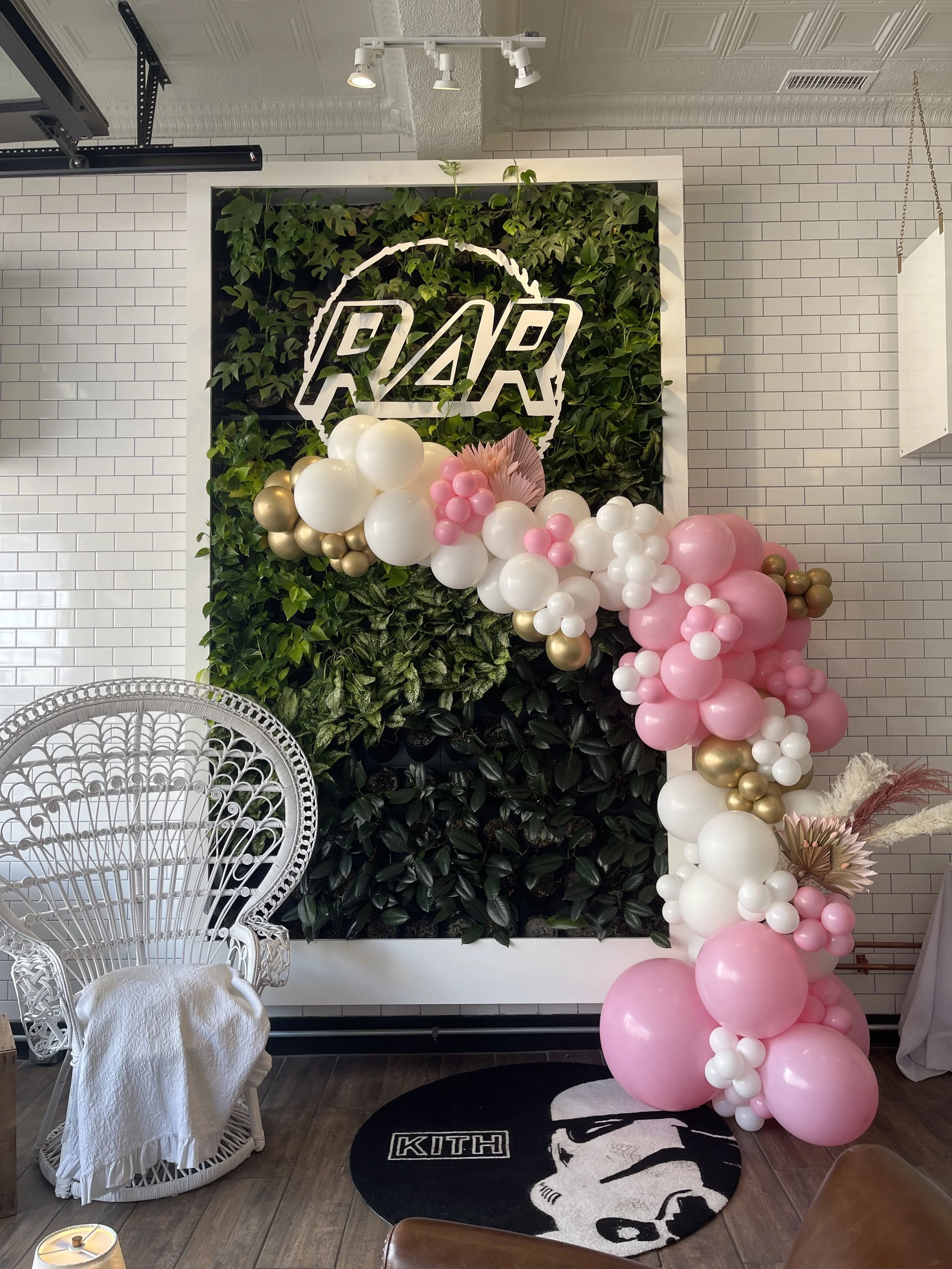 Pink, white, and gold balloon garland framing a custom neon-style 'R&R' sign on a greenery wall