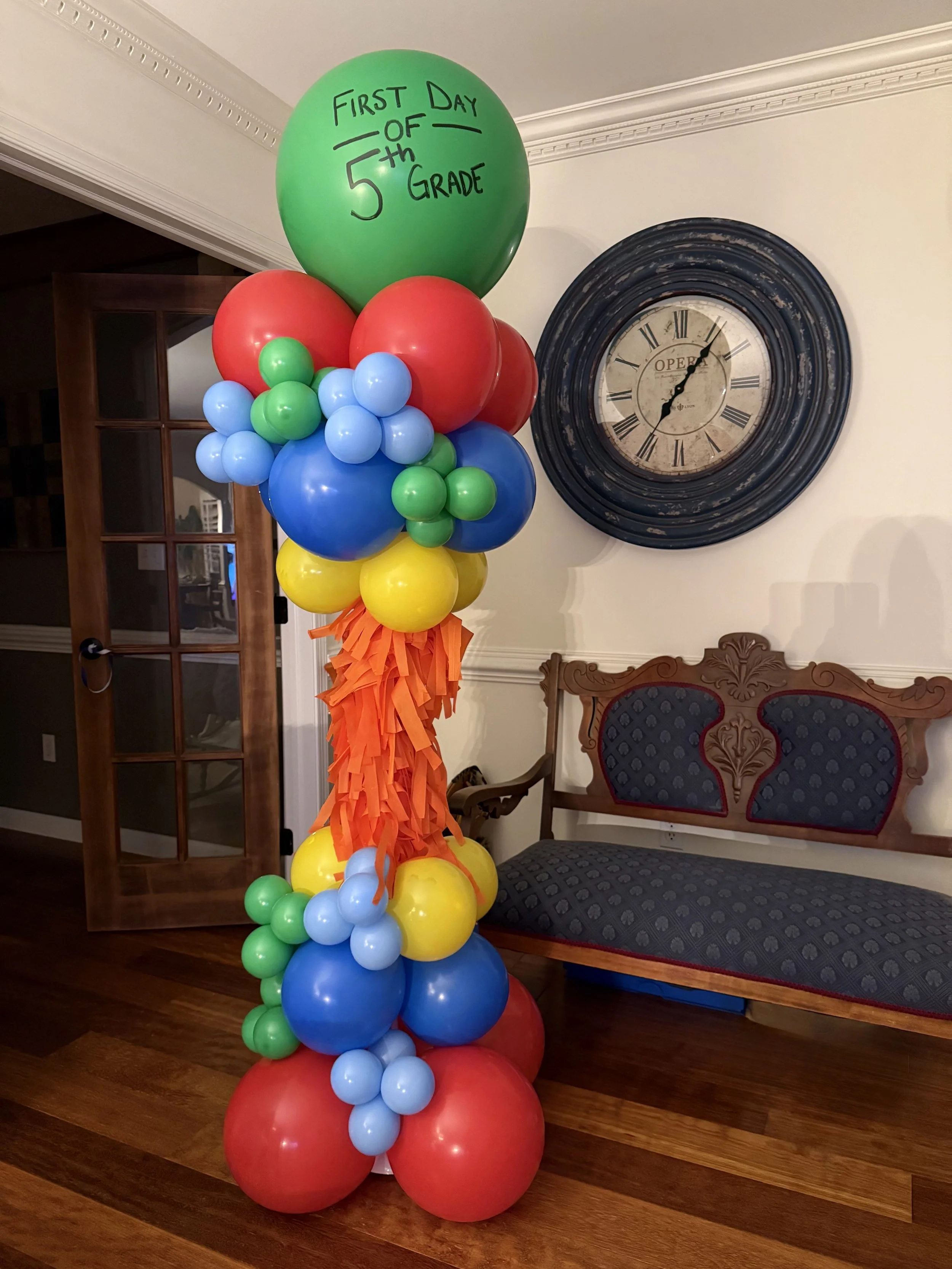 Colorful balloon tower with a green balloon at the top reading 'First Day of 5th Grade' in black marker. The tower includes red, blue, yellow, and green balloons arranged from top to bottom, with orange tissue paper at the base.