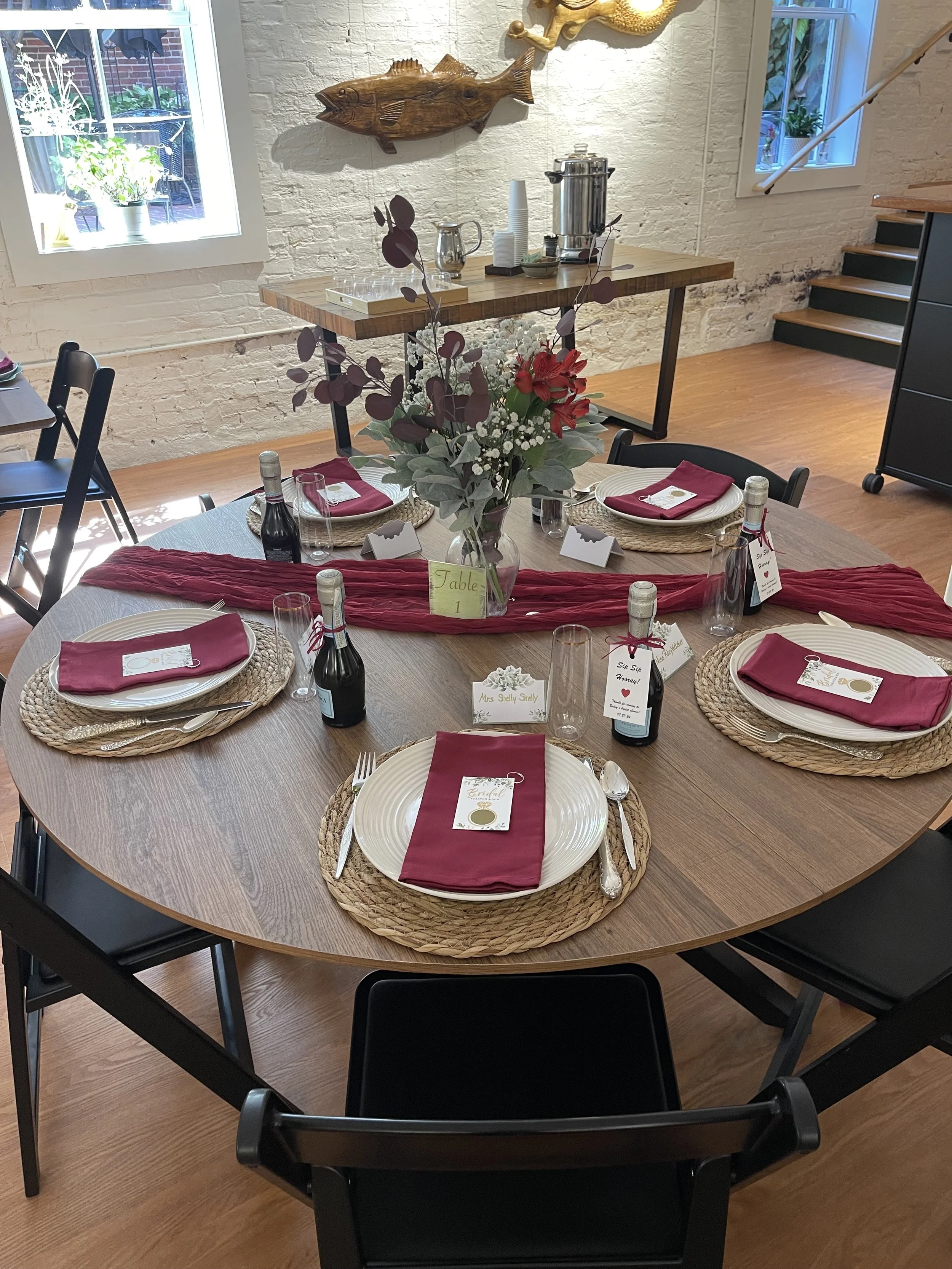 Styled round dinner table with burgundy linens, woven chargers, and a floral centerpiece