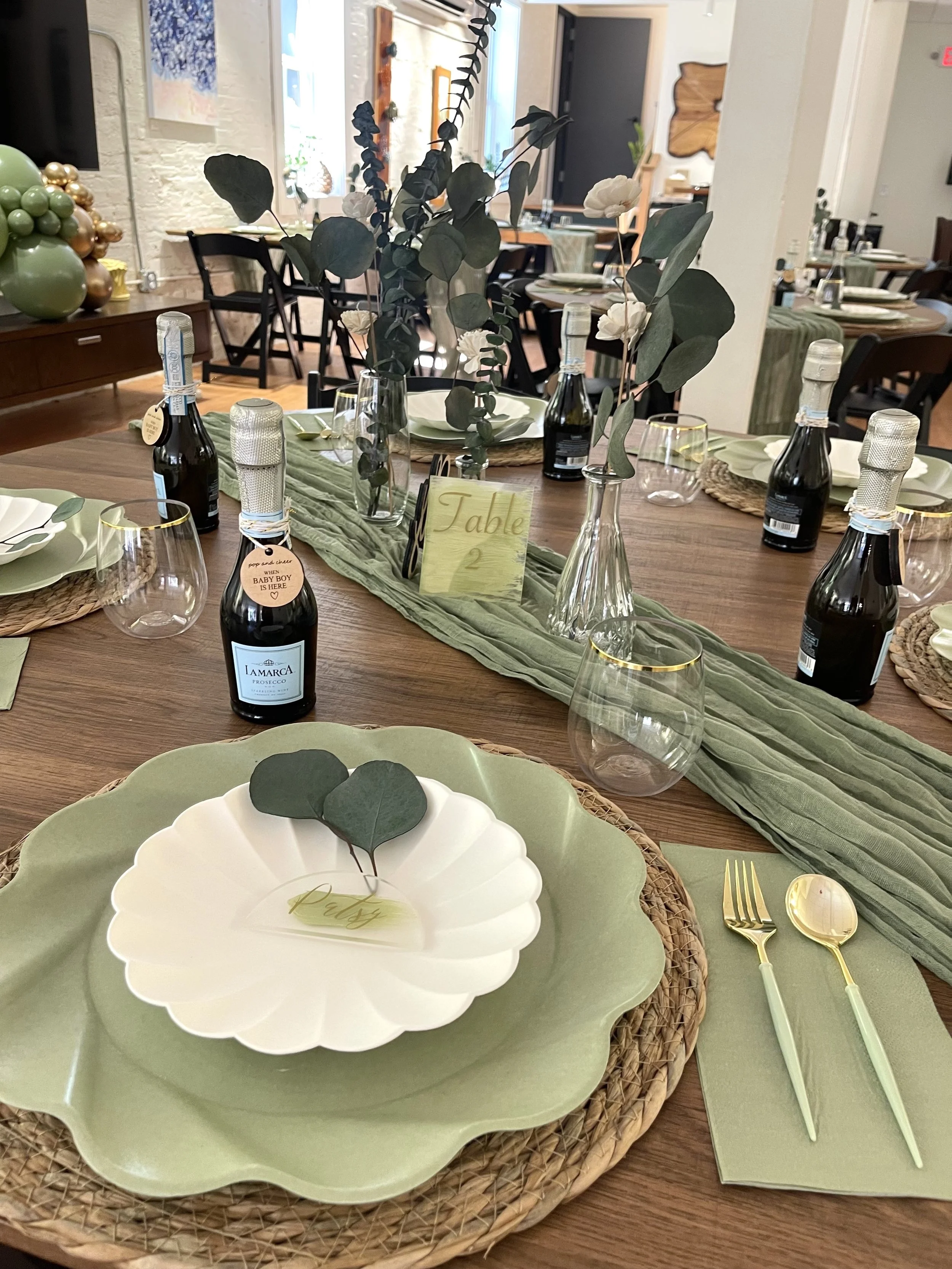 A beautifully set dining table with green and cream-colored plates, gold utensils, wine glasses, a green table runner, and floral centerpieces with eucalyptus leaves and white flowers. There are bottles of sparkling wine with labels, a sign that says 'Table 2,' and additional tables with similar settings in the background.