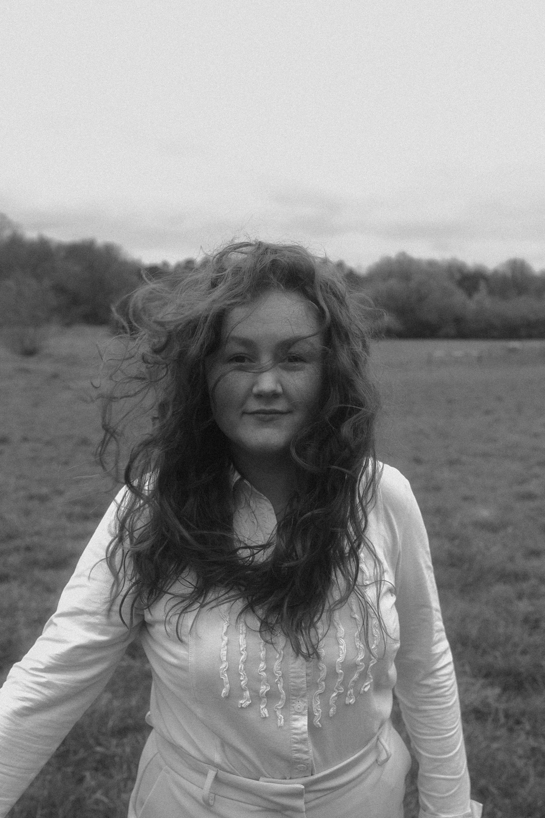 Schwarz-weiß-Foto einer Frau mit langen, gewellten Haaren, die in einem offenen Feld steht, mit Bäumen im Hintergrund, Blick nach rechts oben gerichtet, lächelnd.