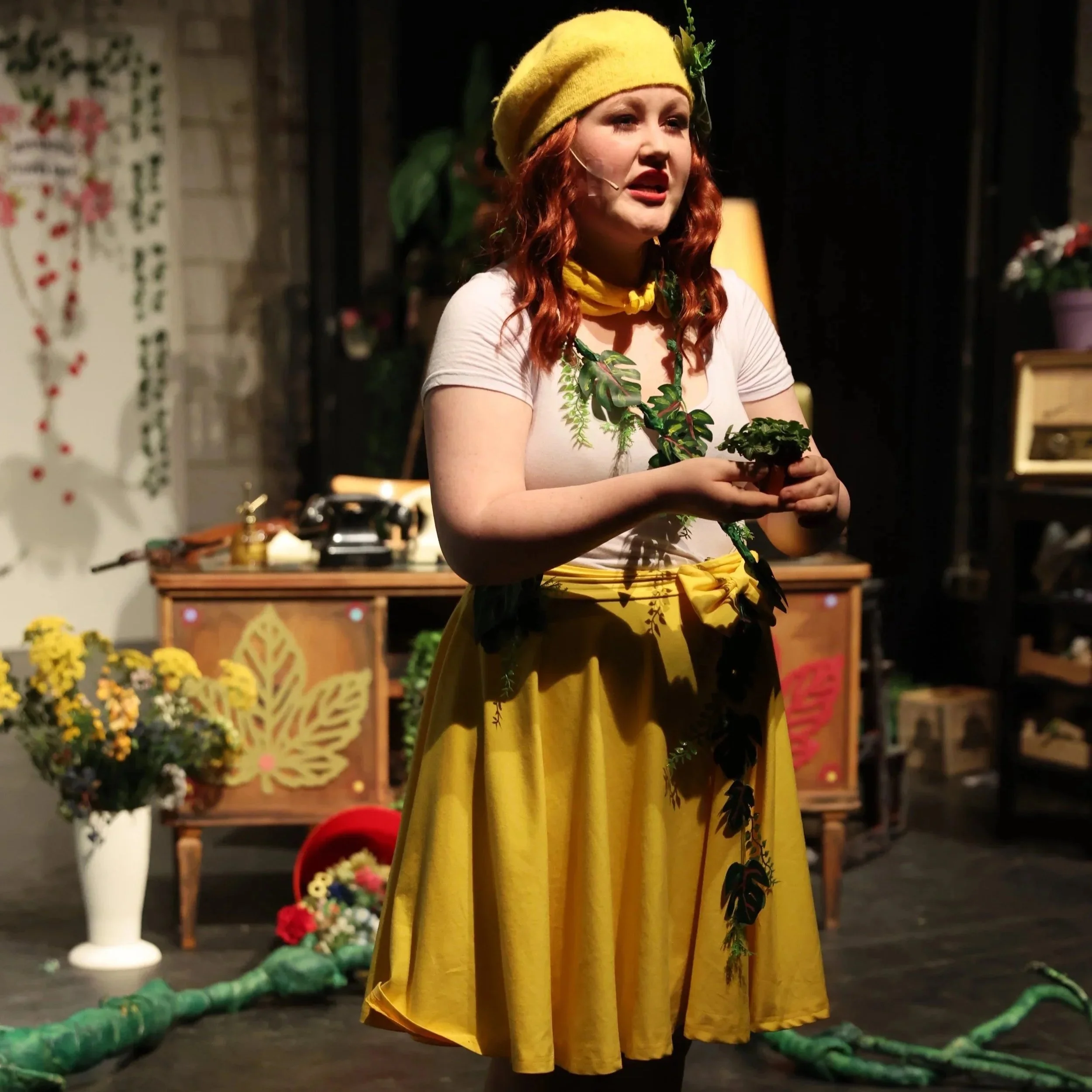 Eine Frau mit roten Locken trägt einen gelben Hut und eine passende gelbe Schürze. Sie steht auf einer Bühne mit dunklem Hintergrund, umgeben von Blumen und Dekorationen, vermutlich bei einer Theateraufführung oder einem Kostümtheater.