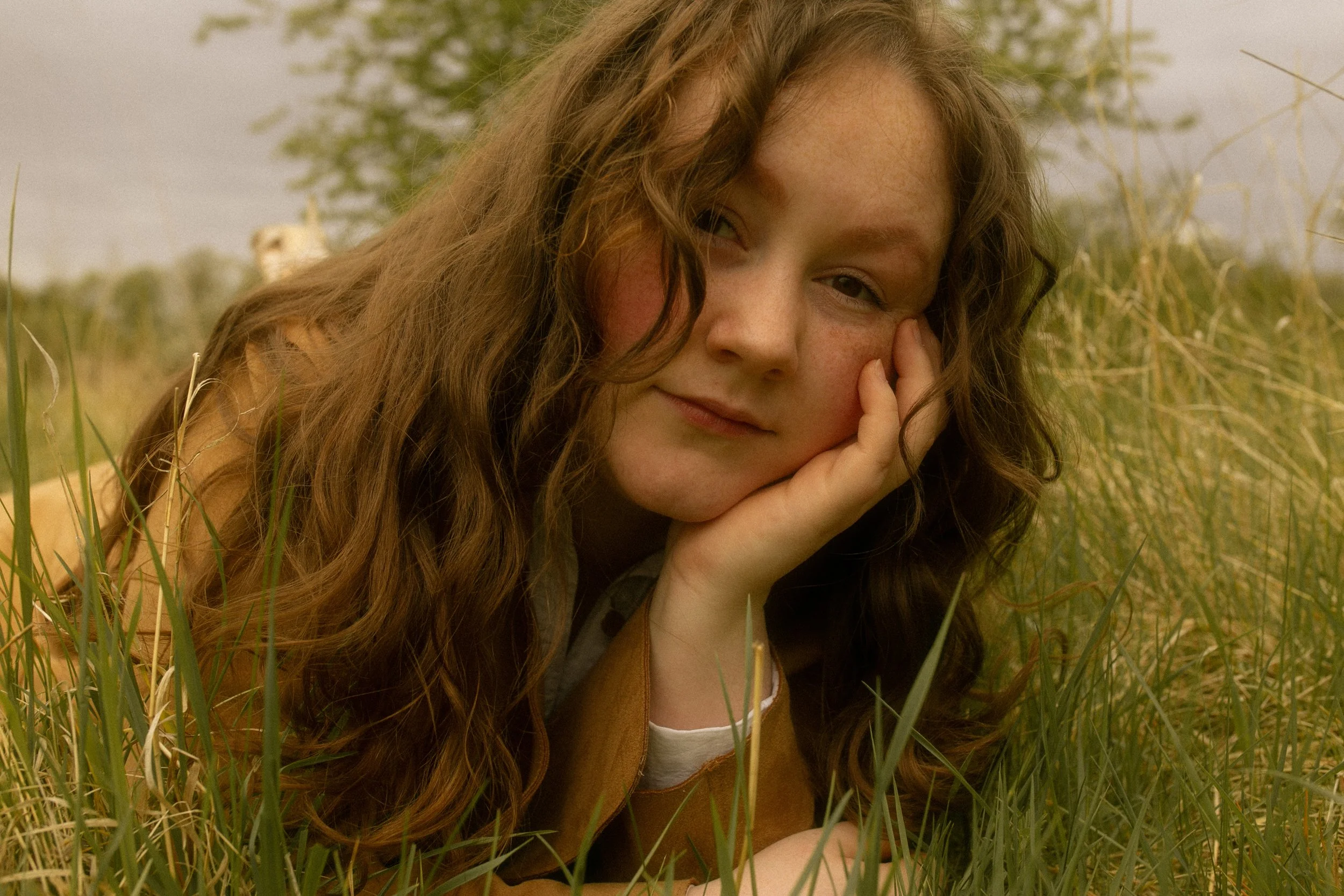 Junge Frau mit longem, lockigem Haar liegt im Gras, berührt ihr Gesicht und blickt zufrieden in die Kamera.
