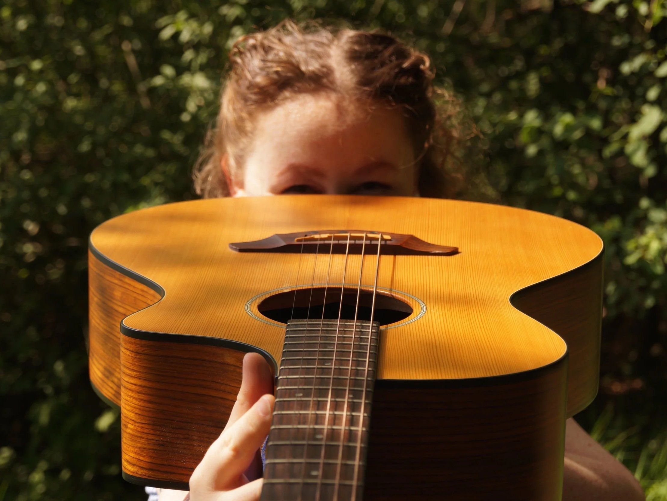 Person spielt Gitarre im Freien mit grünem Laub im Hintergrund.