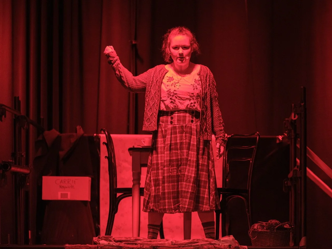 Eine Frau steht auf einer Theaterbühne in einem roten Scheinwerferlicht. Sie trägt einen Rock, ein Oberteil, eine Strickjacke und einen Halsketten. Sie hat eine aggressive Haltung mit erhobenem Arm.