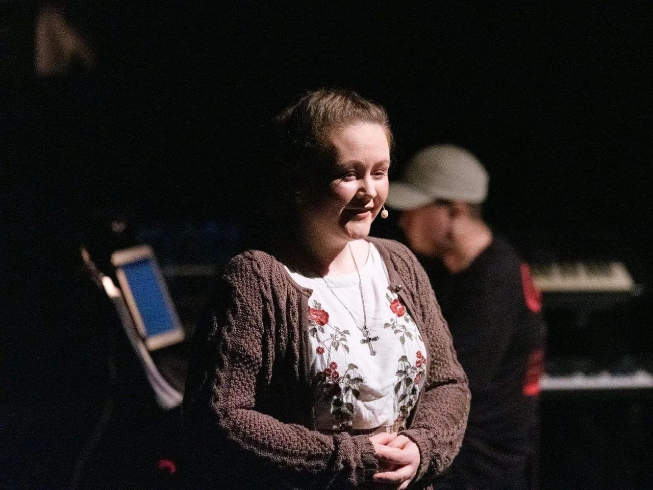 Eine Frau mit einem Mikrofon lächelnd, im Hintergrund eine Person am Klavier, dunkle Kulisse, vermutlich im Theater oder bei einer Veranstaltung.