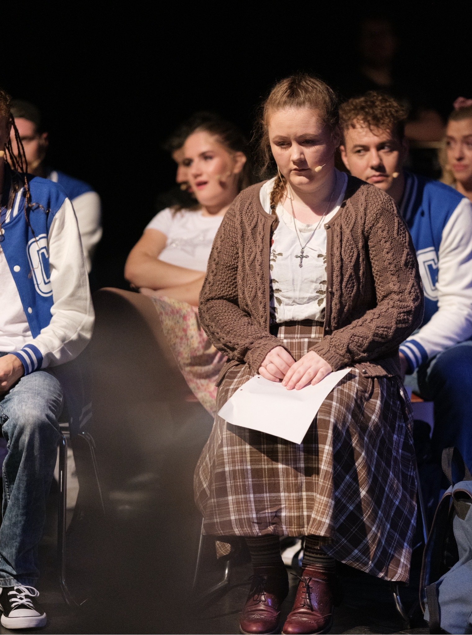 Jugendliche sitzen auf einer Bühne oder in einem Theater, tragen Mikrofone und Jacken, einige halten Papiere in der Hand, im Hintergrund ist dunkler Raum oder Bühne.