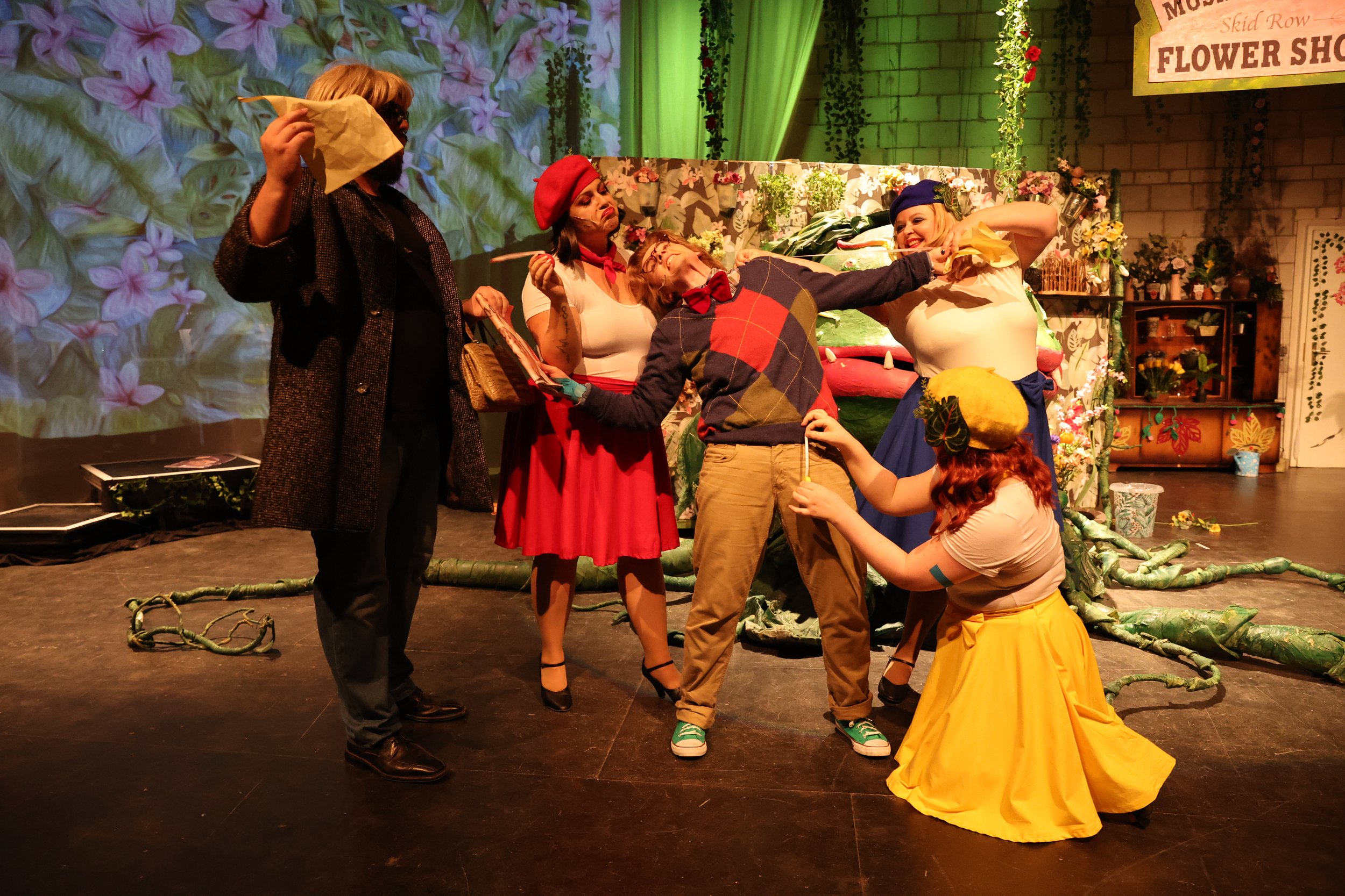 Schauspieler auf Bühne bei Theateraufführung mit Blumen und Dekor in Hintergründen.