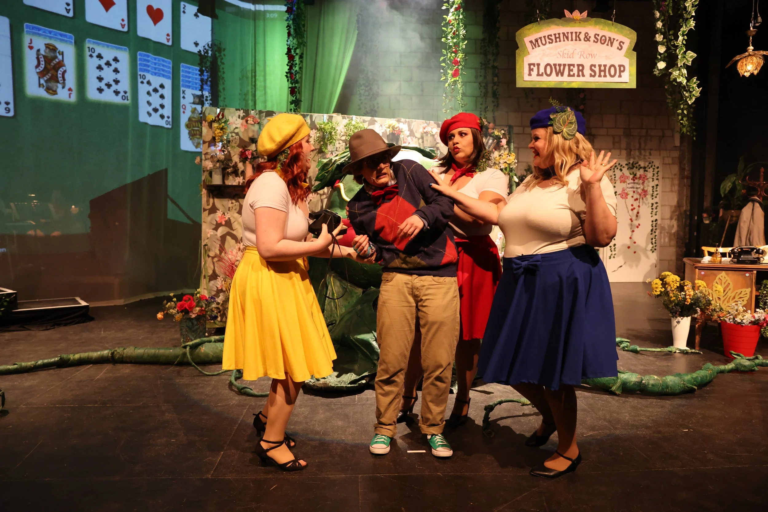 Fünf Schauspieler auf einer Bühne, die in einem Blumenladen spielen, mit einem Schild 'Mushnik & Son's Flower Shop' im Hintergrund. Sie tragen bunte Kleidung und scheinen in einer Szene eines Theaterstücks zu agieren.