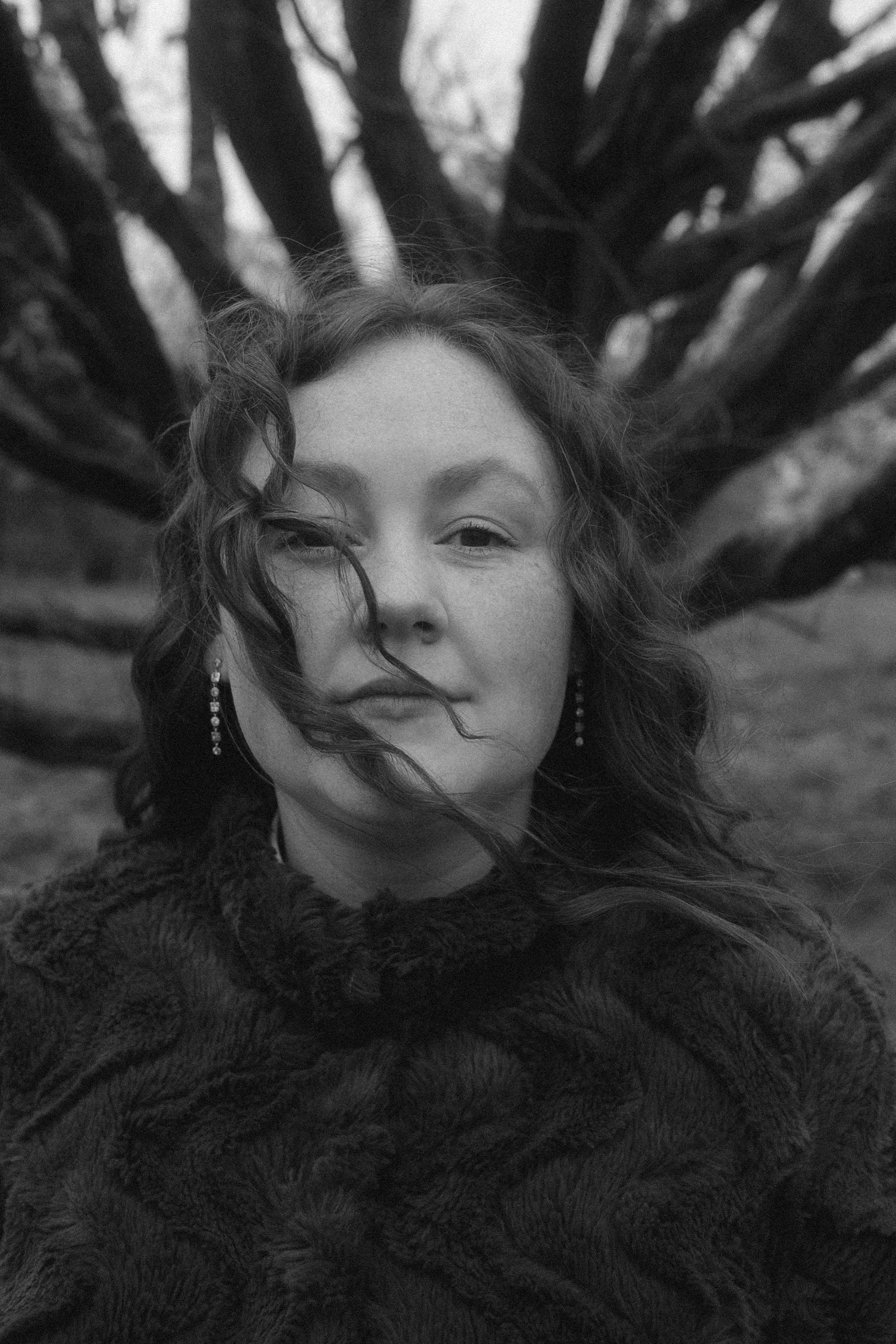 Schwarz-weiß-Foto eines Frauenkopfes, Nahaufnahme, mit schwarzen Locken, trägt Ohrringe, im Hintergrund ein Baum mit verzweigten Ästen.