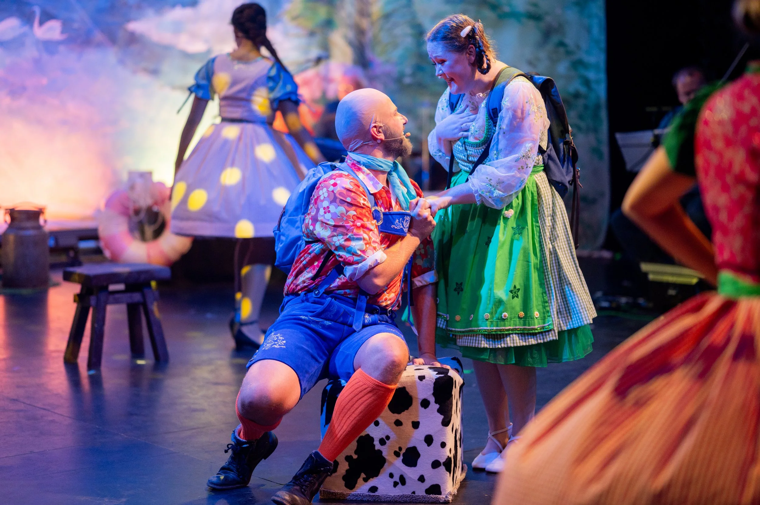 Menschen in farbenfrohen Kostümen auf einer Theaterbühne, eine Person kniet mit einem blumenmusternden Rock und trägt einen Mikrofon, während eine andere Person mit einer Umarmung oder Unterstützung spricht.