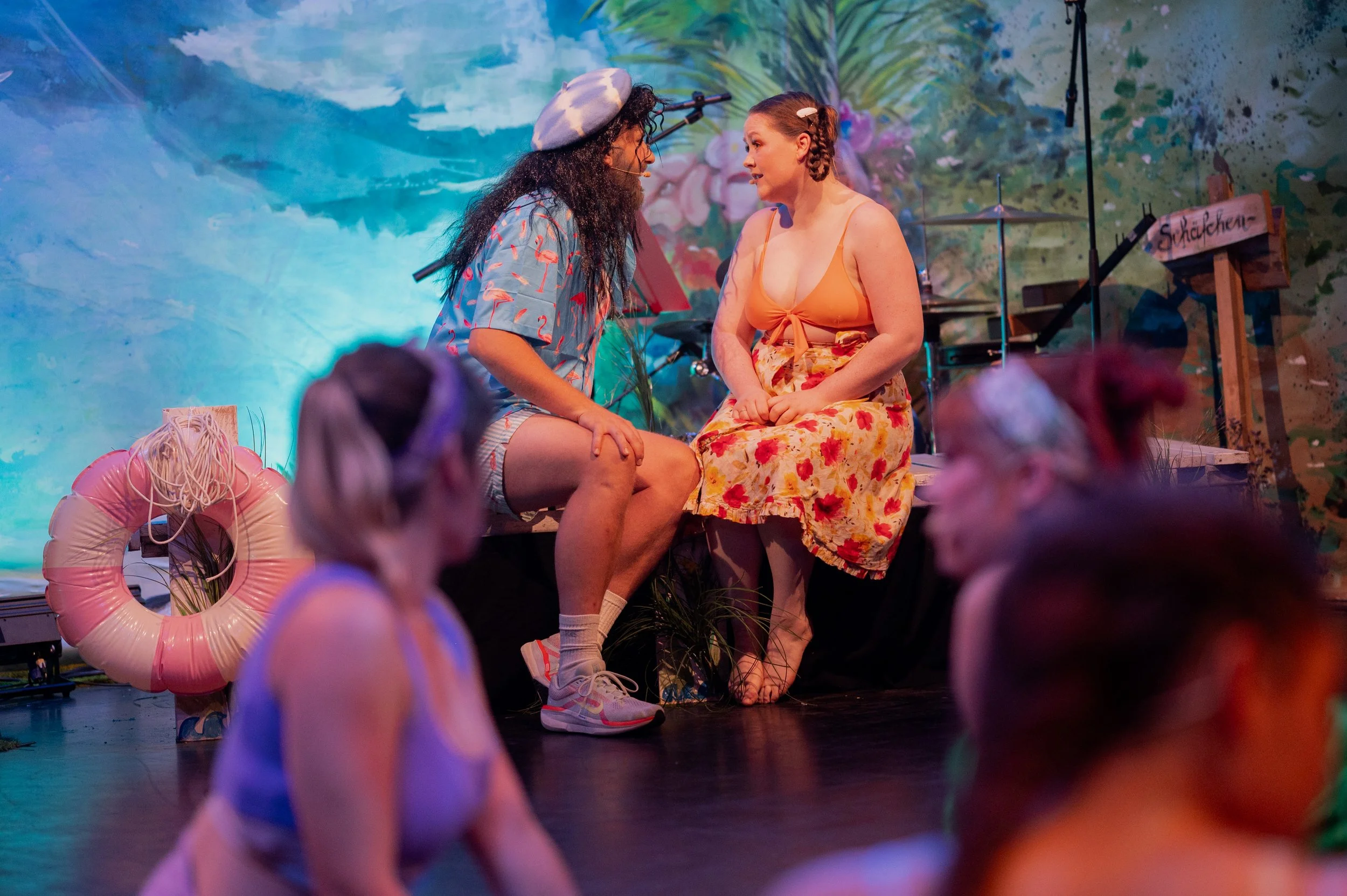Zwei Frauen sitzen sich bei einer Theateraufführung gegenüber, im Hintergrund eine bunte Wand mit Pflanzenmotiven und Musikinstrumenten.