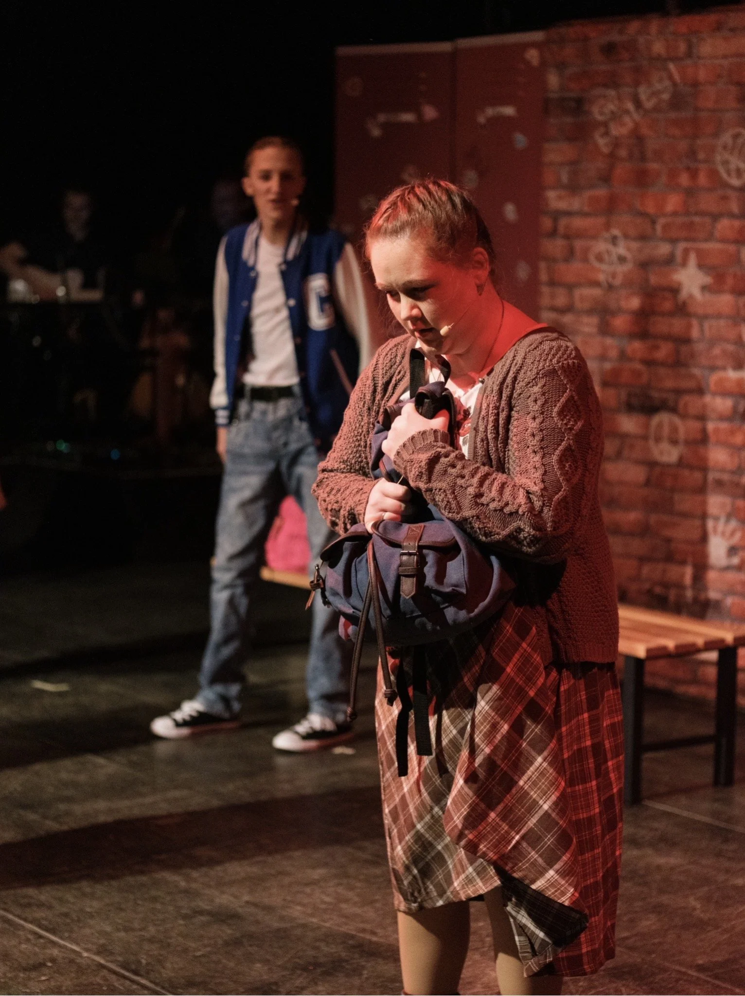 Eine junge Frau steht mit einem Mikrofon in der Hand auf einer Bühne, schaut nach unten, während im Hintergrund eine andere junge Frau und eine Person mit Gitarre auf einer dunklen Theater- oder Bühnenumgebung sind.