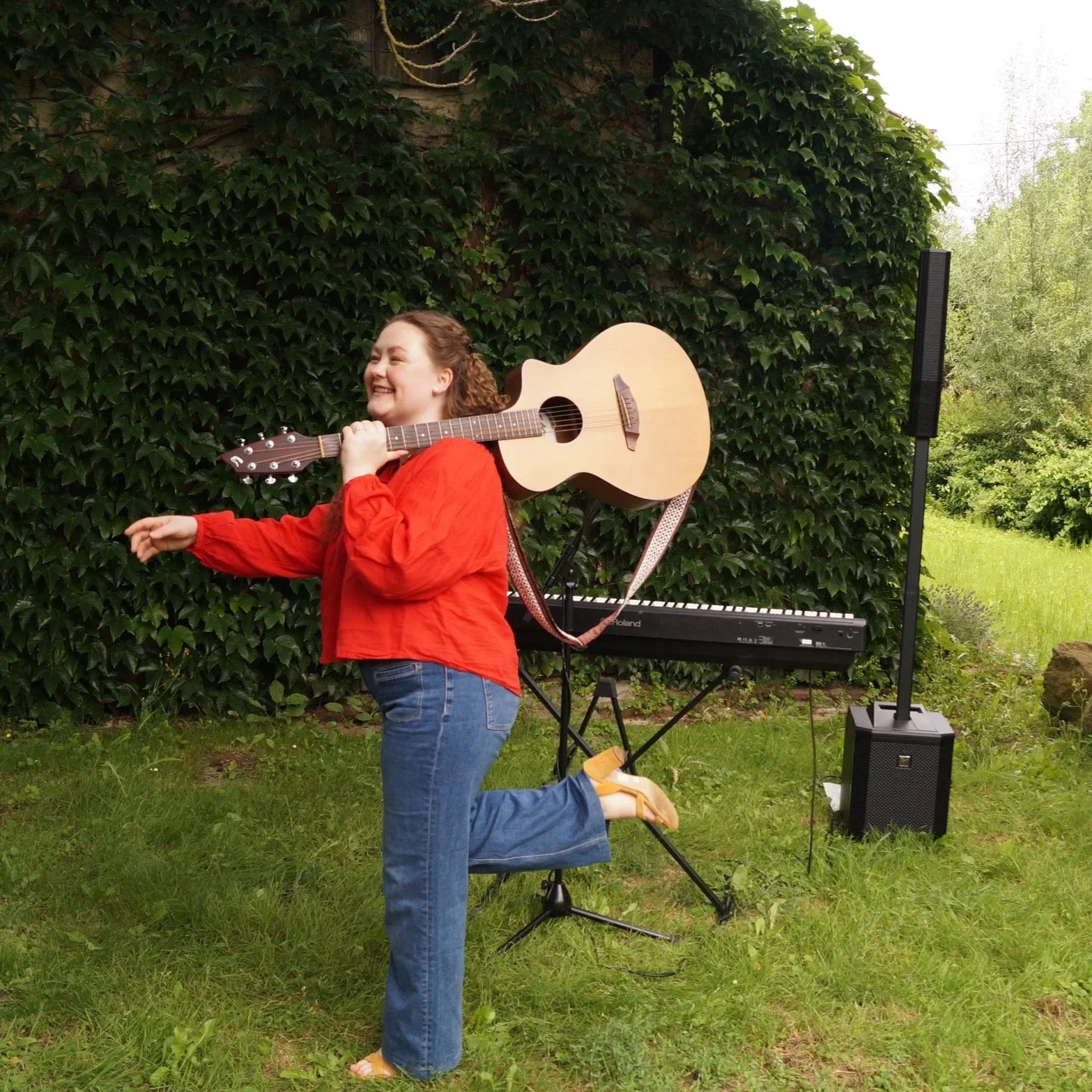 Eine junge Frau mit lockigem Haar, die in einem Garten lacht, während sie ein Akustikgitarre auf den Schultern trägt. Im Hintergrund steht ein Keyboard und ein Lautsprecher auf grünem Rasen, umgeben von grünen Büschen und Bäumen.