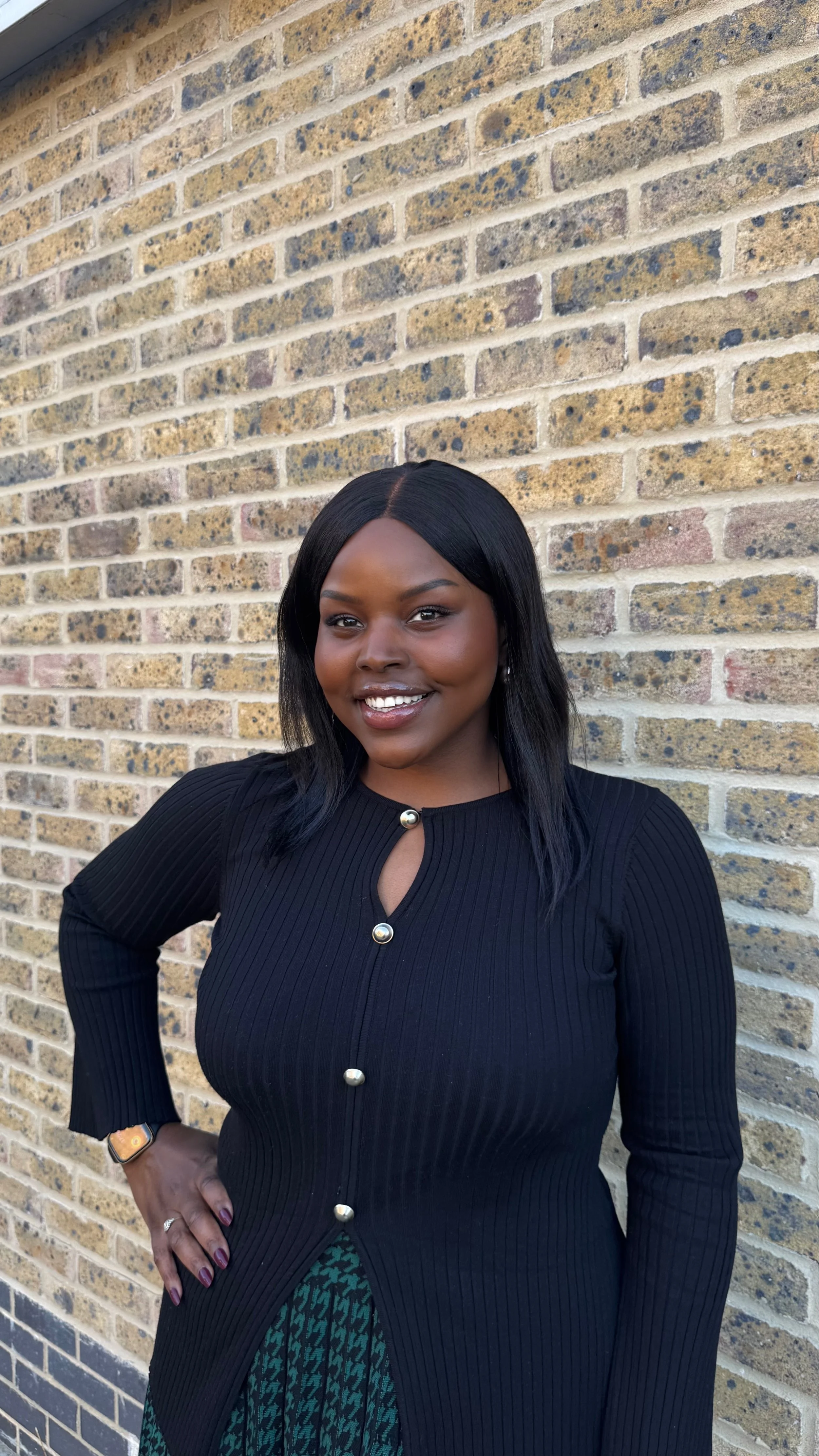 A woman with dark hair and dark skin, smiling, standing in front of a brick wall, wearing a black ribbed cardigan with pearl buttons and a patterned skirt.